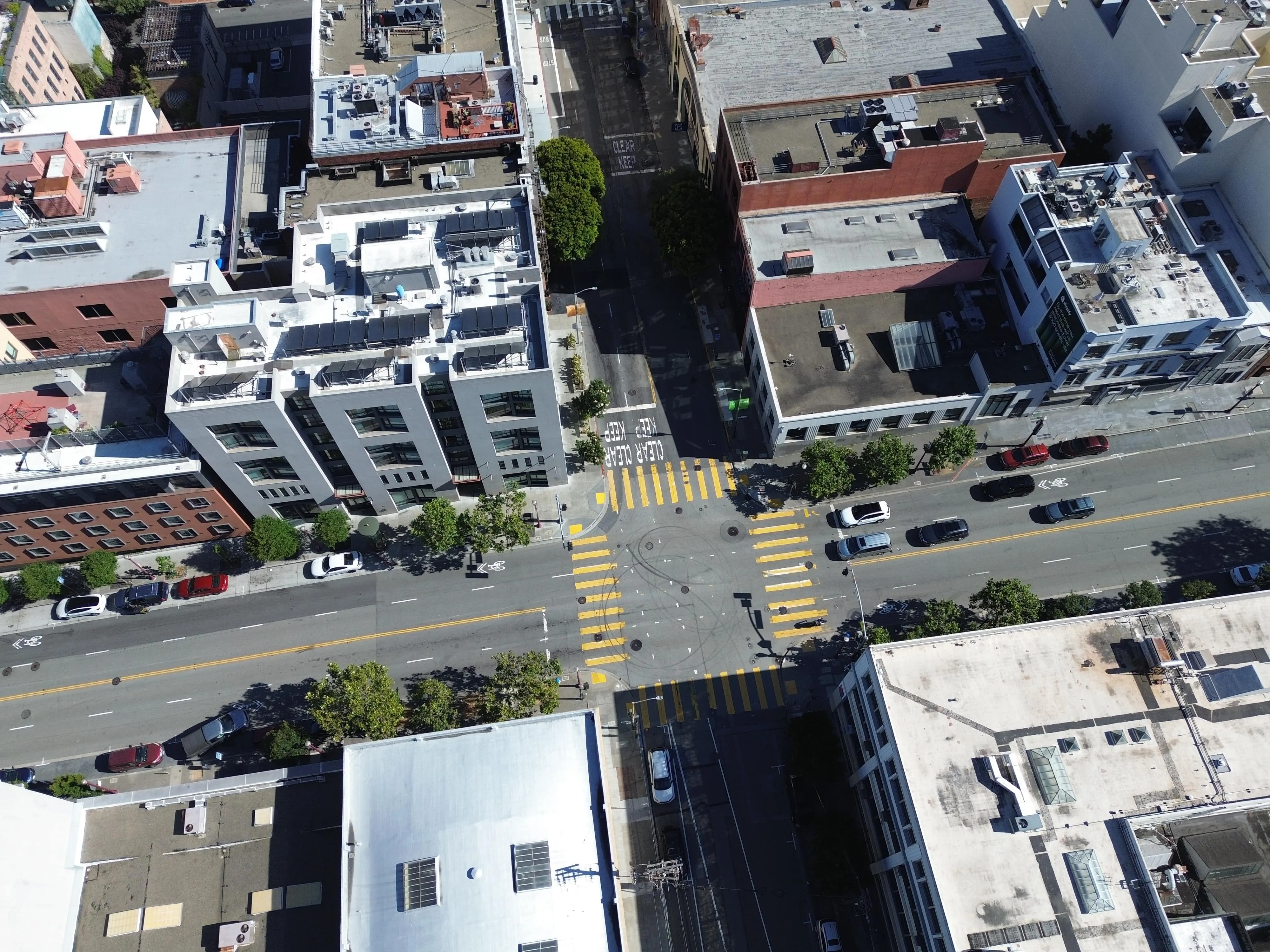 Aerial view of an urban street intersection with yellow crosswalk lines and several parked cars beside multi-story buildings.