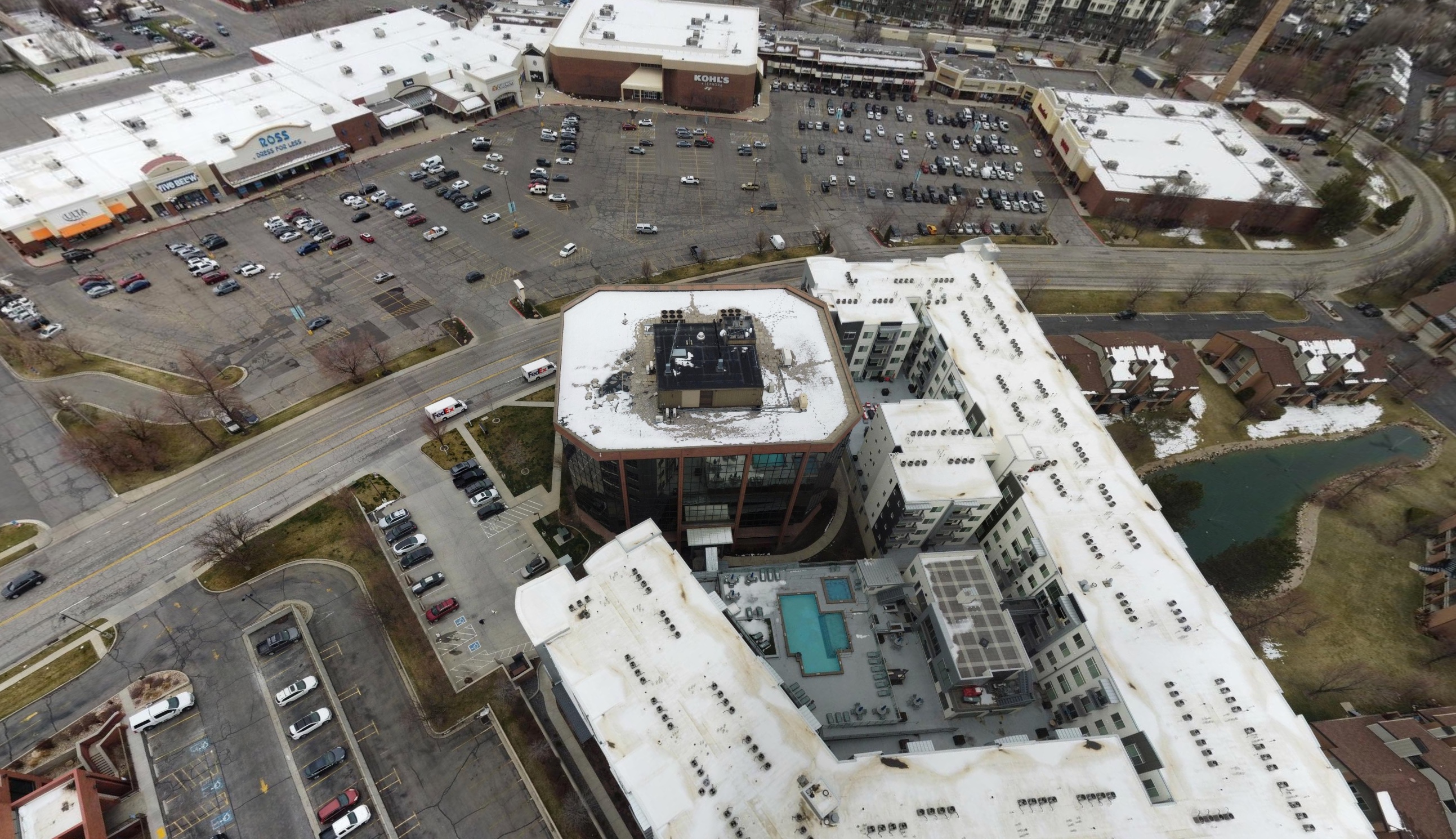 Aerial view of a shopping center parking lot near a large apartment complex with a swimming pool and snow on rooftops.