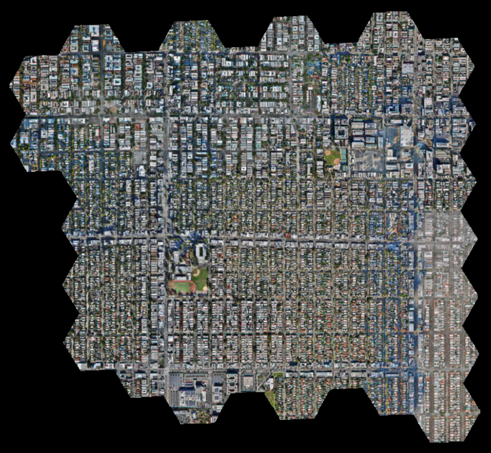 Aerial view of a densely built urban area with a grid-like street pattern, including residential and commercial buildings, and small green spaces with sports fields.