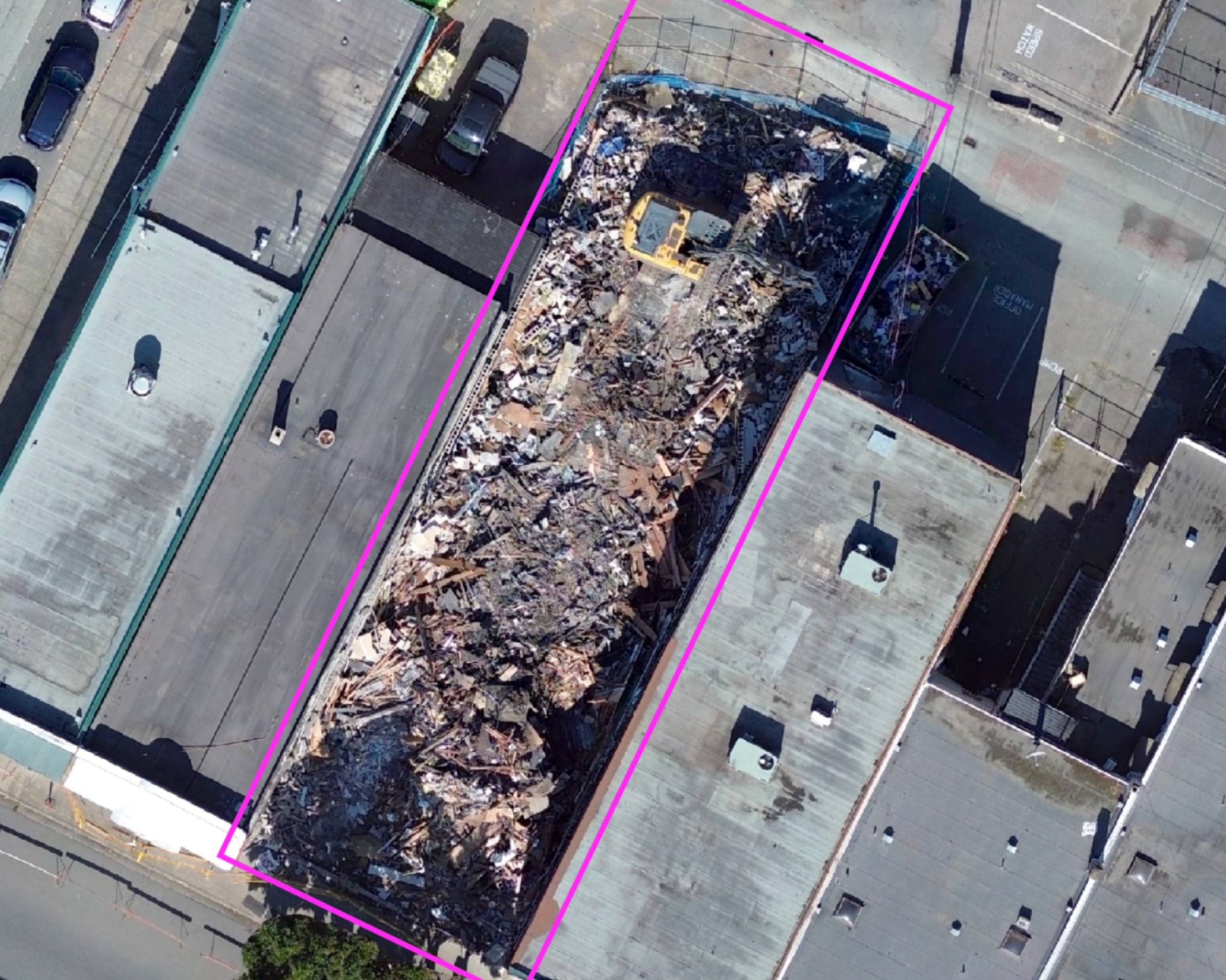Aerial view of a demolished building site marked with a magenta boundary, surrounded by intact rooftops and parked cars.