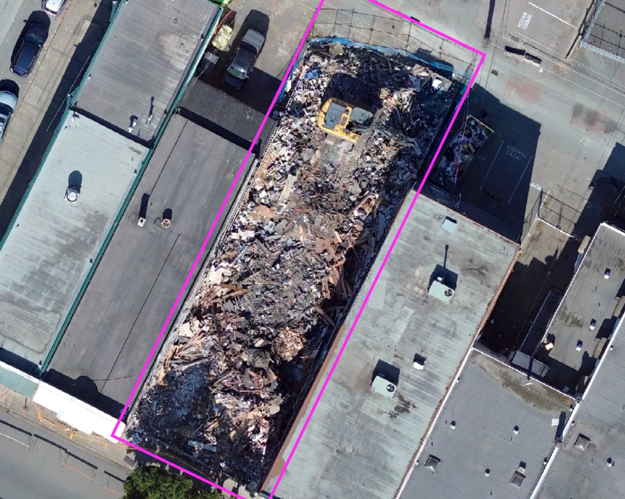 Aerial view of a demolished building site marked with a magenta boundary, surrounded by intact rooftops and parked cars.