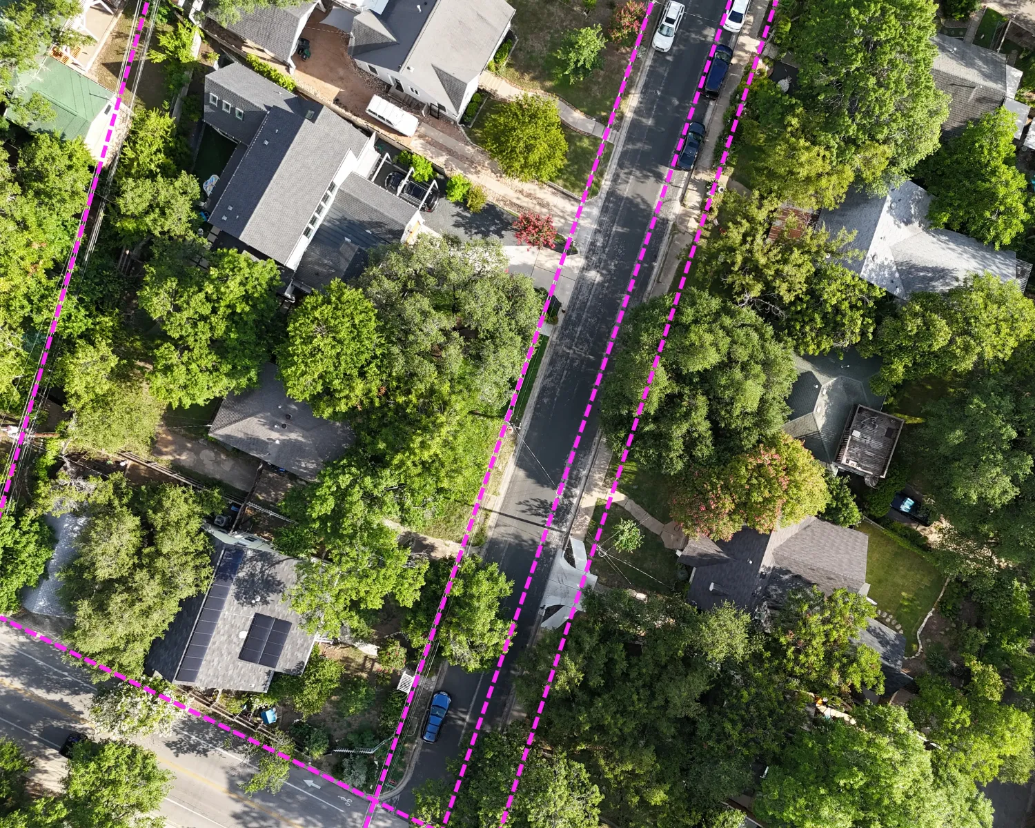 Aerial view of a residential neighborhood with houses, trees, parked cars, and magenta dashed lines overlaying property boundaries.