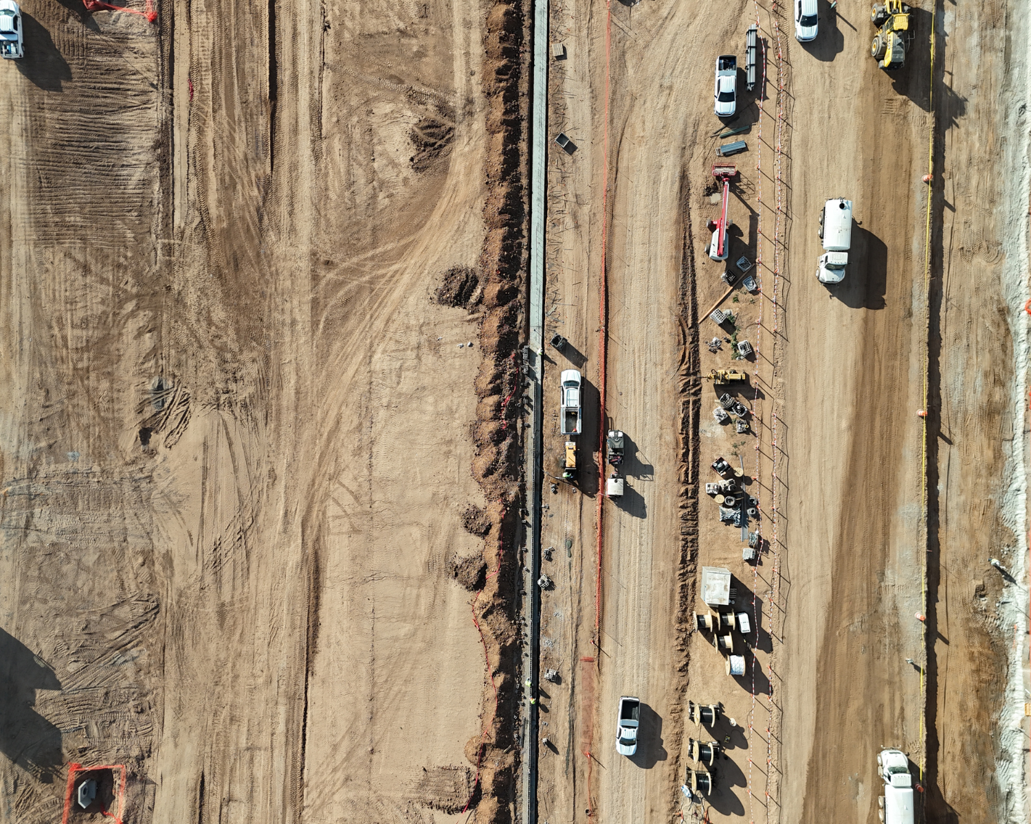 Aerial view of a construction site with dirt ground, several white vehicles, construction equipment, and materials lined up along a dirt road.