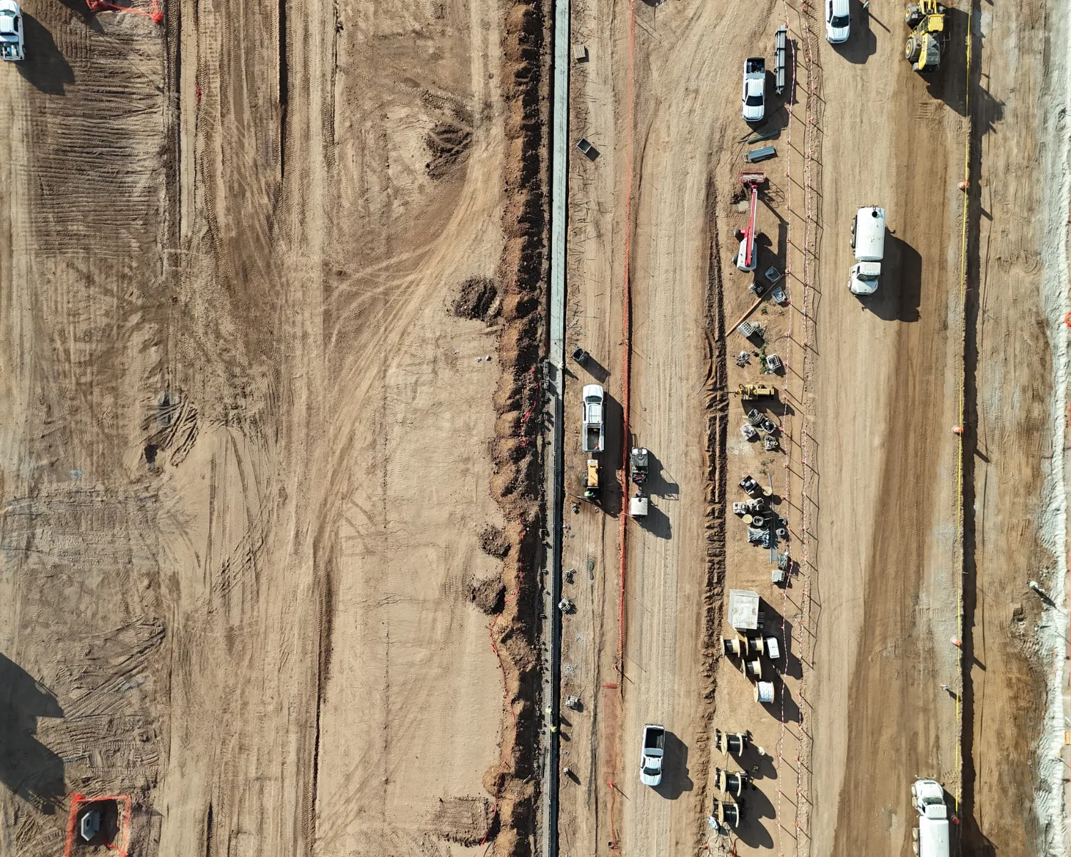 Aerial view of a construction site with dirt ground, several white vehicles, construction equipment, and materials lined up along a dirt road.