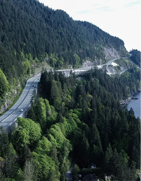 Curving highway along a dense forested mountainside next to a large body of water.
