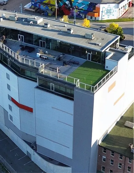 Modern building rooftop with glass railings, lounge chairs, tables, and green turf area.