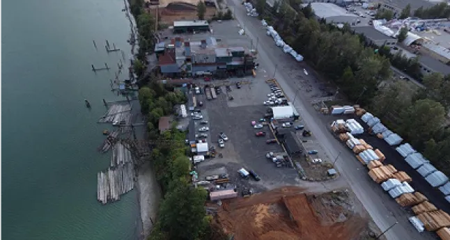 Aerial view of industrial yard with parked vehicles, buildings, stacks of lumber, and adjacent water body.