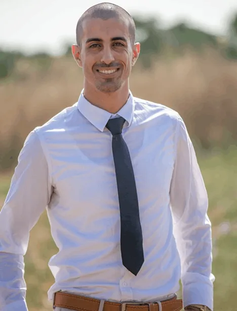 Smiling man in a white shirt and dark tie standing outdoors with a blurred natural background.
