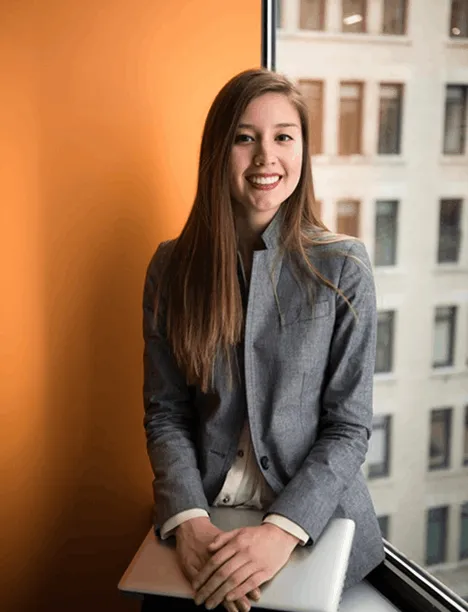 Young woman in gray blazer smiling and holding a laptop while sitting by a window with building view.