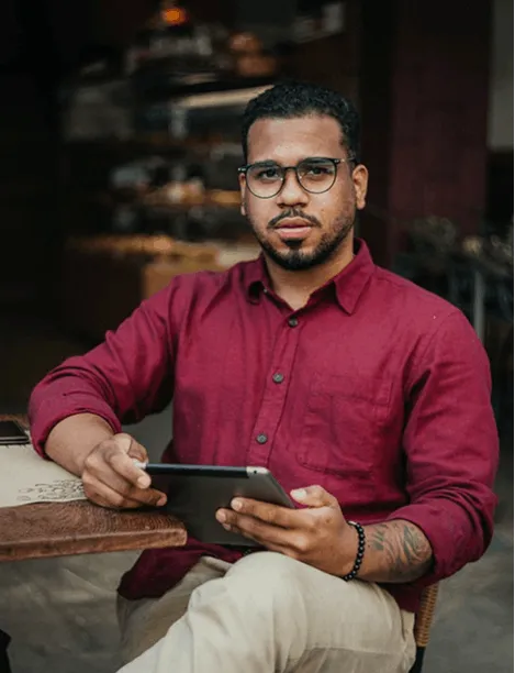 Man with glasses in a red shirt holding a tablet, seated at a table in a cafe.