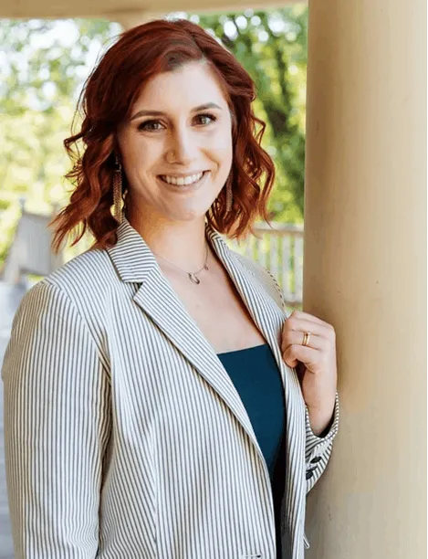 Smiling woman with curly red hair wearing a striped blazer, standing by a beige pillar outside.