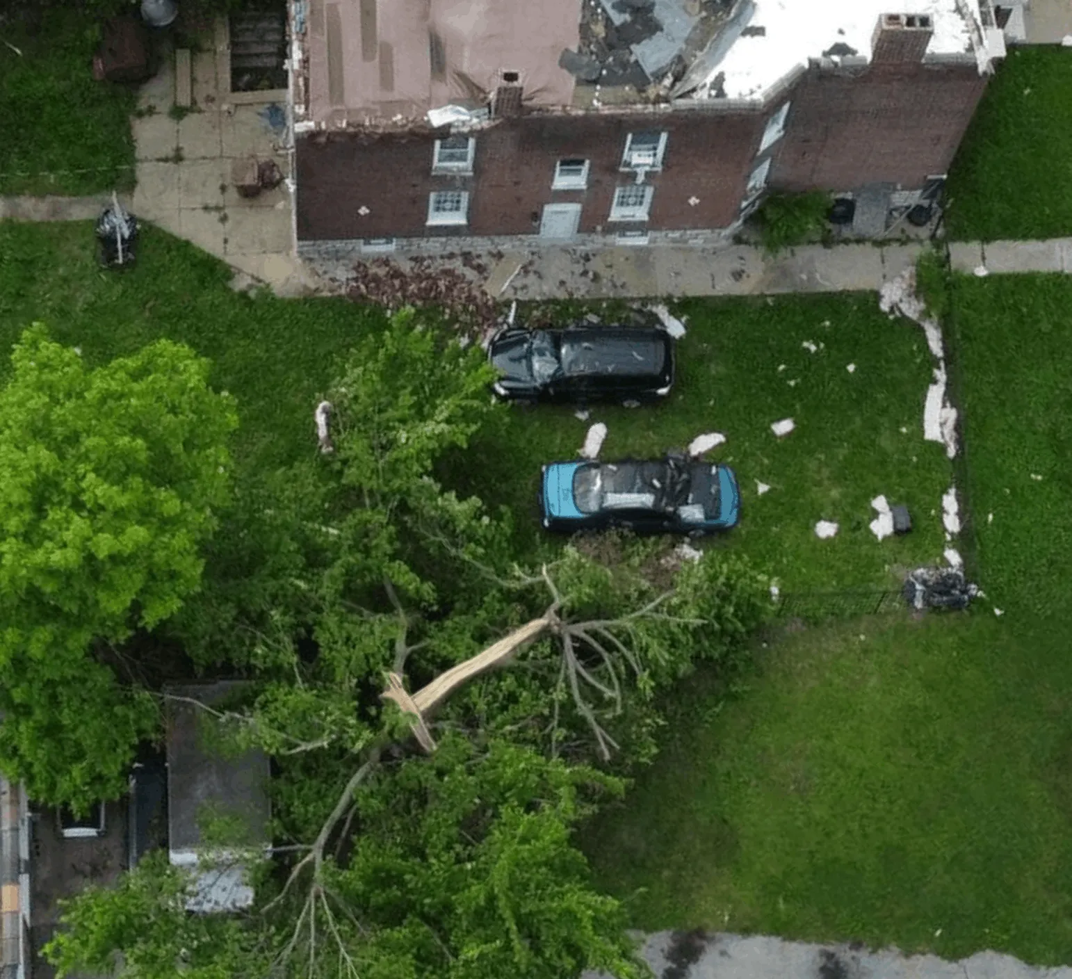 A fallen tree rests on two damaged cars behind a brick house with debris scattered around.