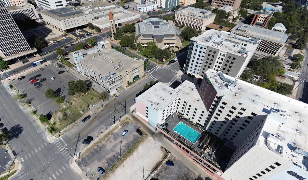 Aerial view of a city block with multi-story buildings, parking lots, streets, and a blue pool.