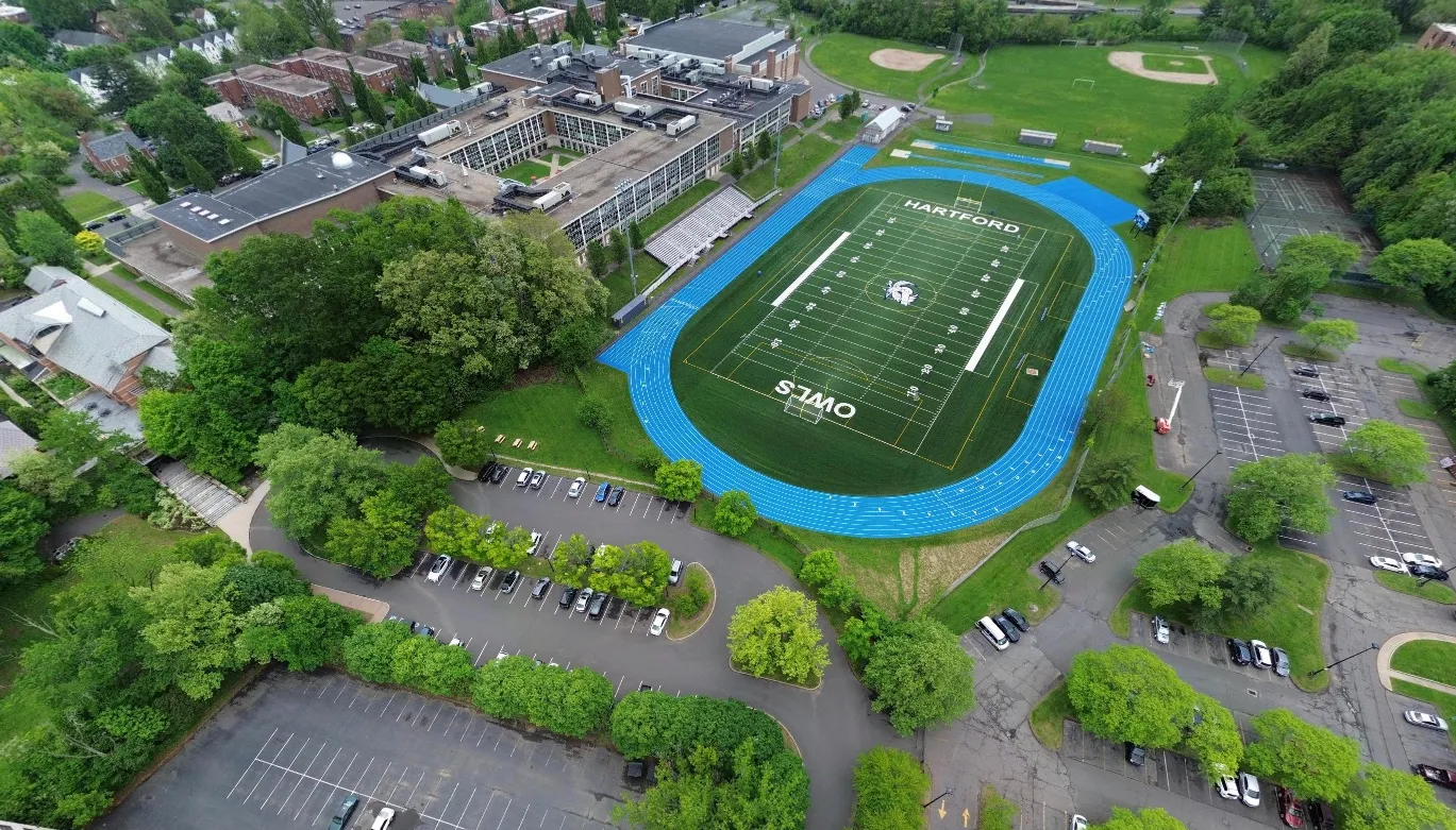 Aerial view of a school with a football field, blue running track, parking lots, and green trees.