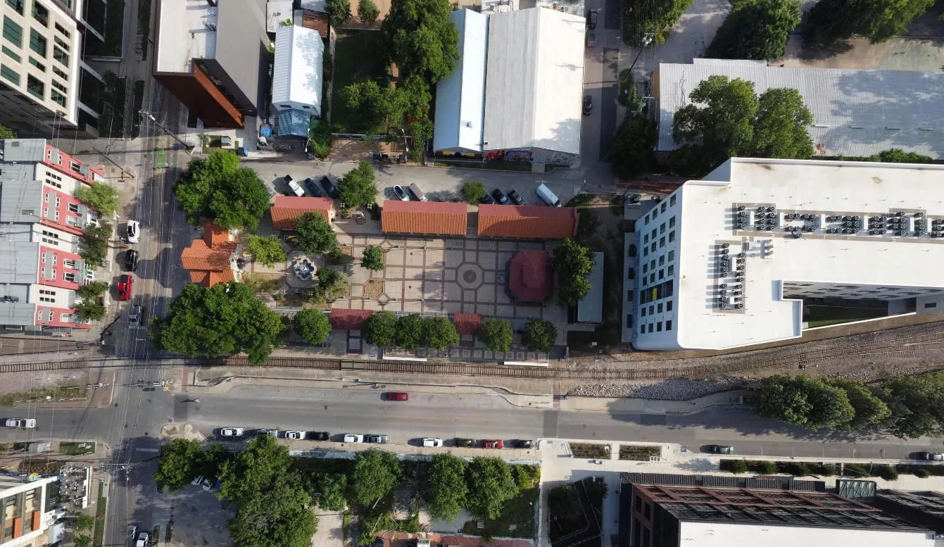 Aerial view of urban park with fountain, trees, surrounding buildings, railroad, and parked cars.