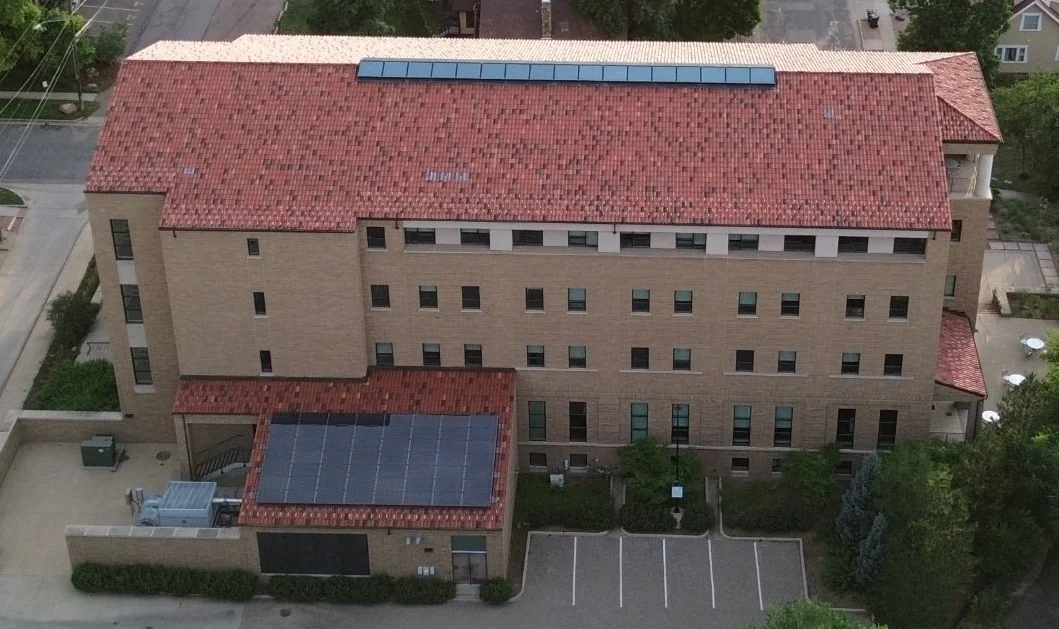 Oblique capture of the building's sandstone facade: rows of windows, the red tile roof and skylight strip, and the solar-panel wing above the parking lot.