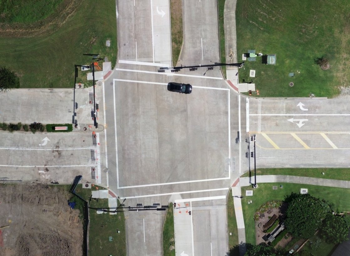 Orthomosaic of a road intersection, viewed straight down: lane arrows, crosswalk lines, traffic signal poles, curbs and landscaping are all map-ready and distinct.