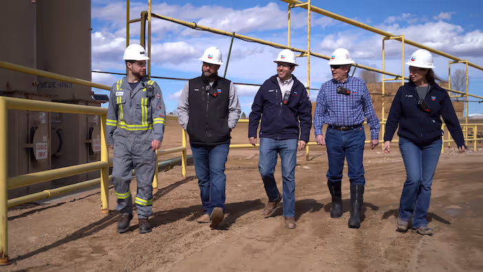Five Chevron workers wearing helmets and safety gear walking on dirt path at industrial site.