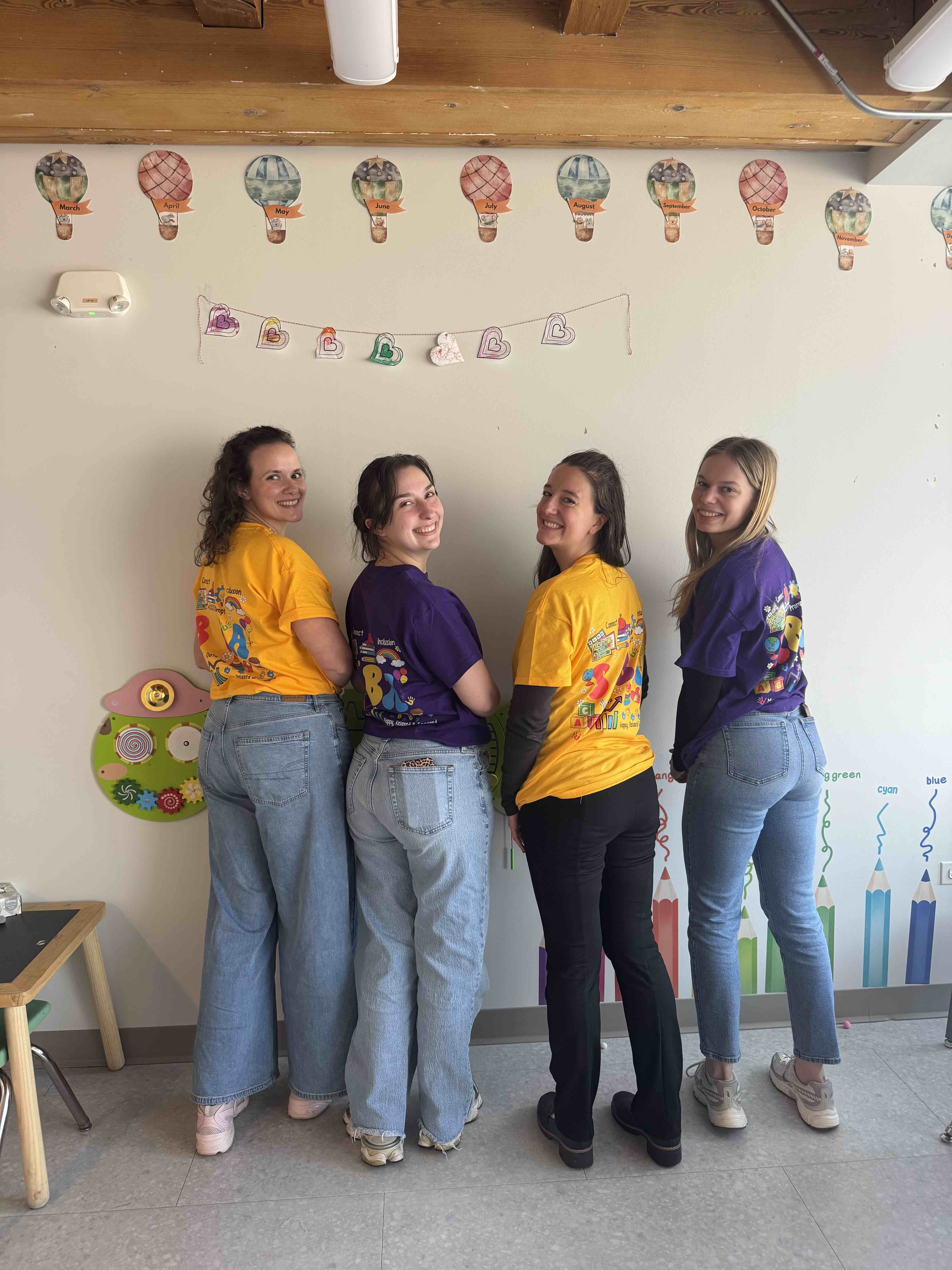 Four women smiling, standing back-to-back wearing colorful graphic T-shirts in a decorated classroom.