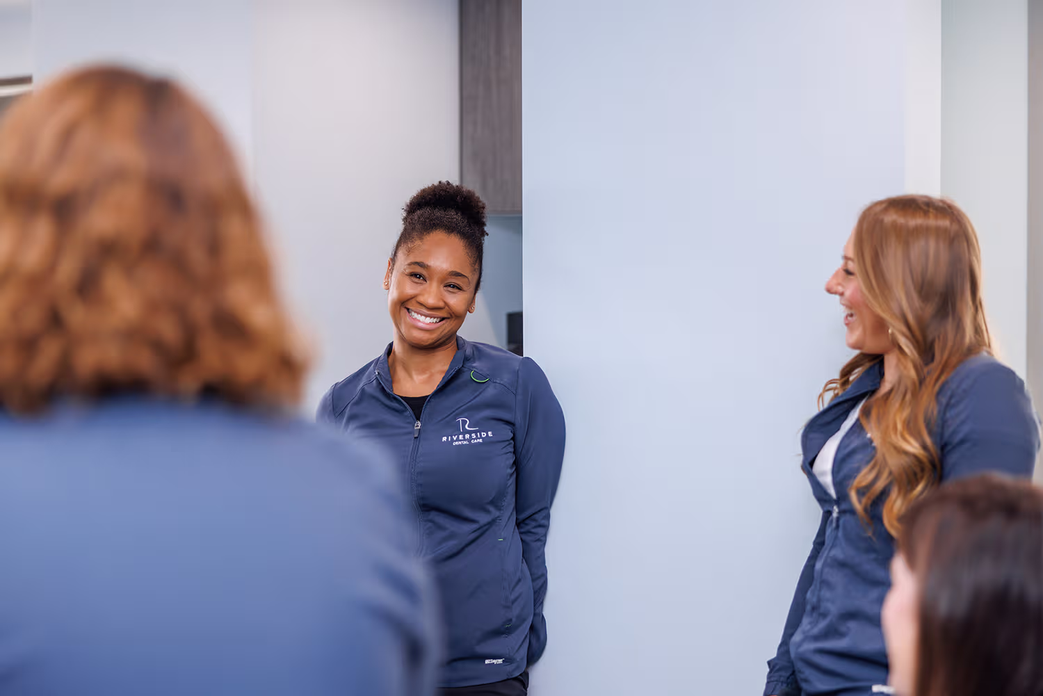 Three women in navy blue Riverside Dental Care jackets smiling and interacting indoors.