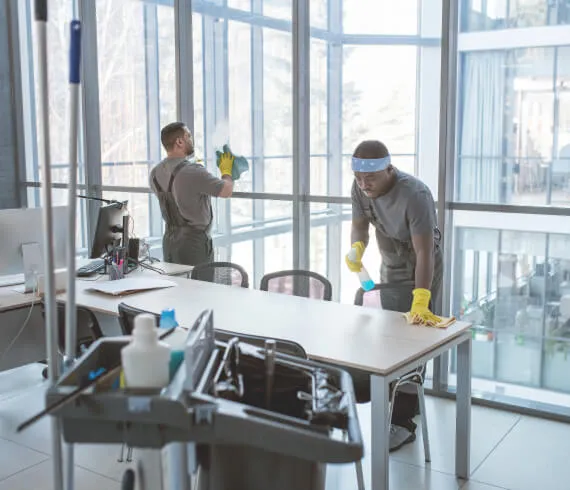 Two professional cleaners wearing gloves cleaning a modern office with large windows and desks.