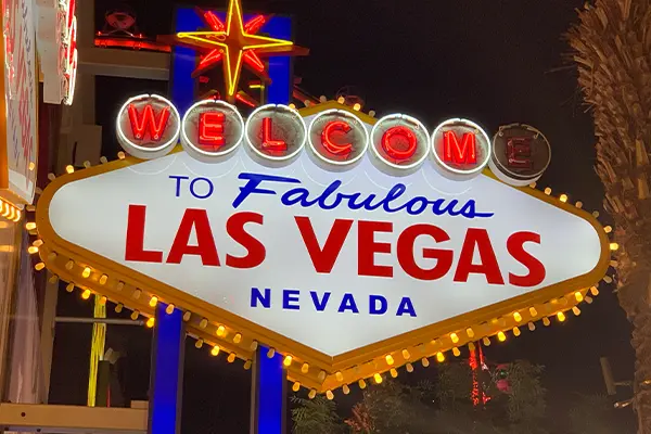 The "Welcome to Fabulous Las Vegas Nevada" neon sign is illuminated at night. The sign features a starburst at the top, red and yellow lights along the edge, and is framed by a palm tree on the right side.