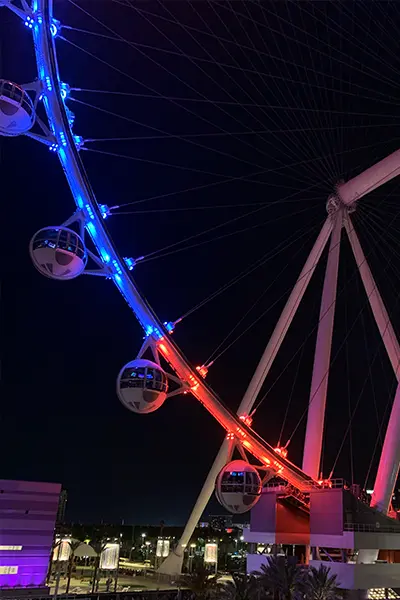 A nighttime view of the High Roller ferris wheel in Las Vegas with its spokes illuminated in blue and orange-red lights. Several passenger capsules are visible on the wheel, which is set against the night sky.