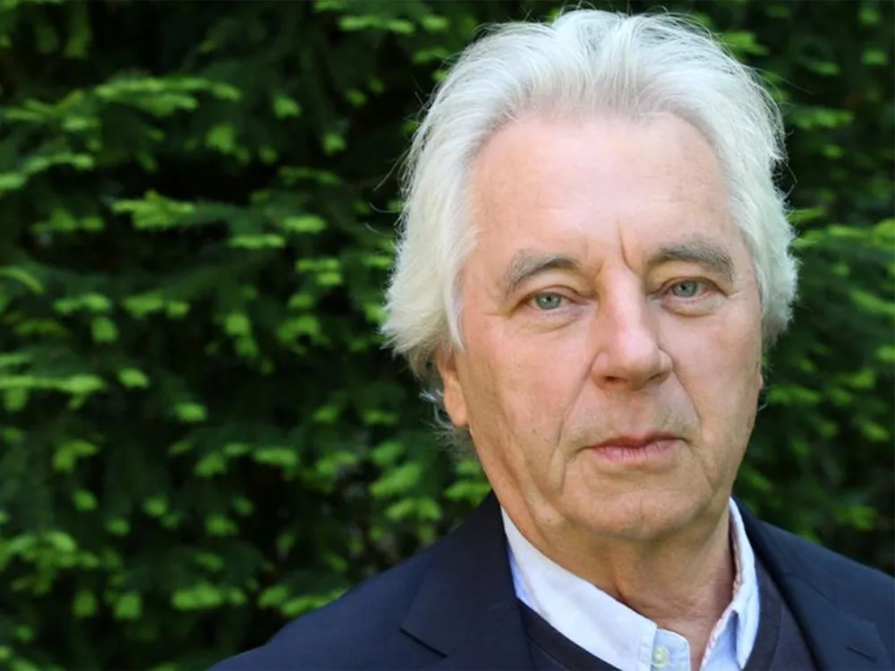 Older man with white hair in a dark suit in front of green foliage outdoors.