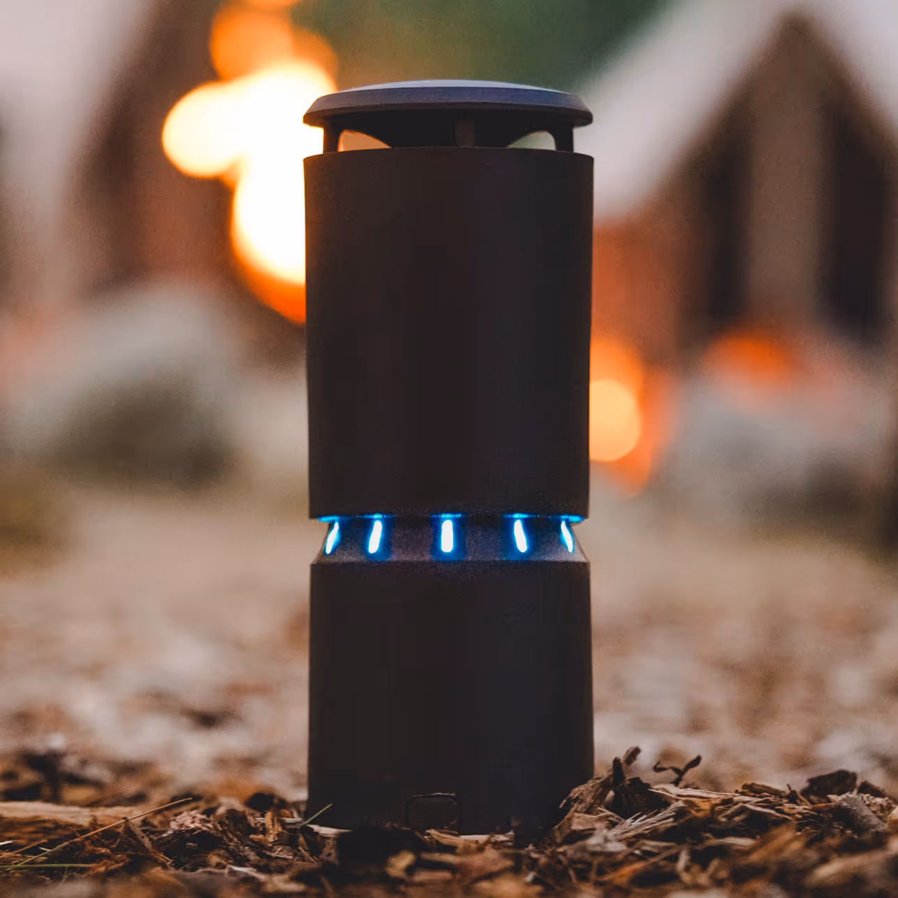 Black cylindrical outdoor mosquito trap with blue LED lights glowing near the middle, placed on bark mulch with a blurred fire in the background.