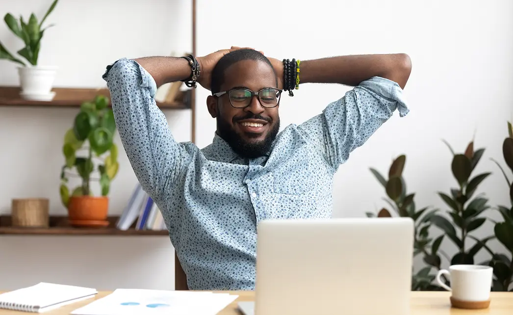 Smiling man with glasses and beard leaning back with hands behind head at desk with laptop and cup.