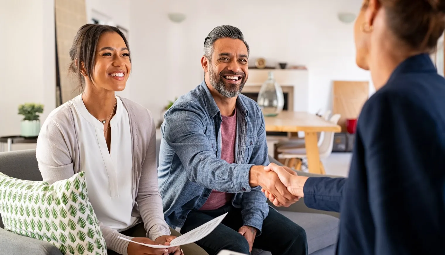 Smiling couple sitting on couch shaking hands with a person in a business setting.