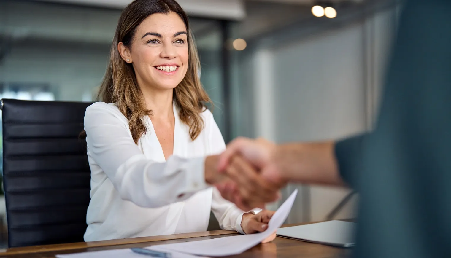 Smiling woman in white blouse shaking hands with a person in an office setting.