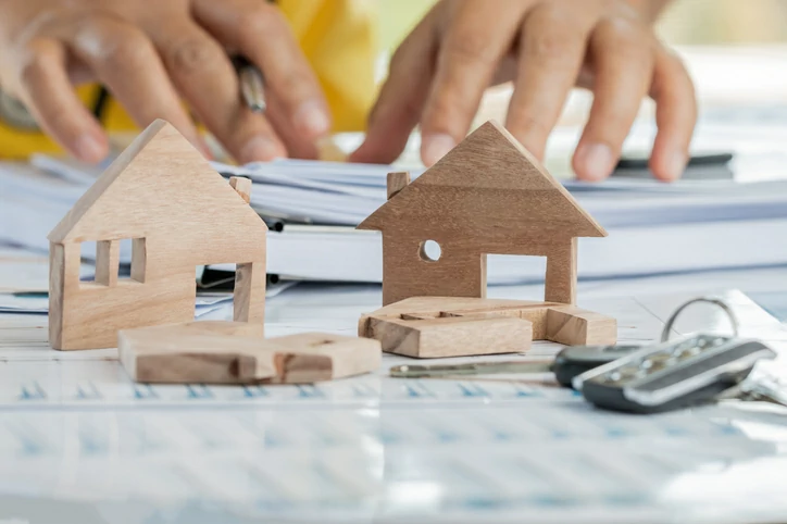 petite maison en bois, clé, calculatrice et main tenant un stylo en arrière plan.