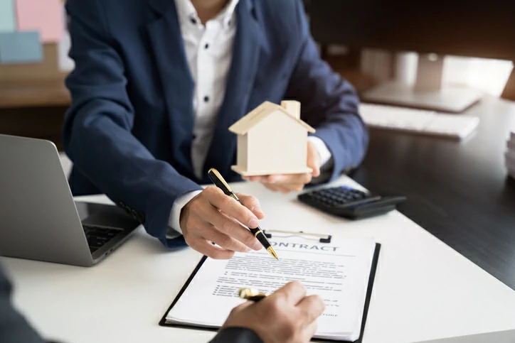 main tenant une petite maison et un stylo et une autre main signant un document