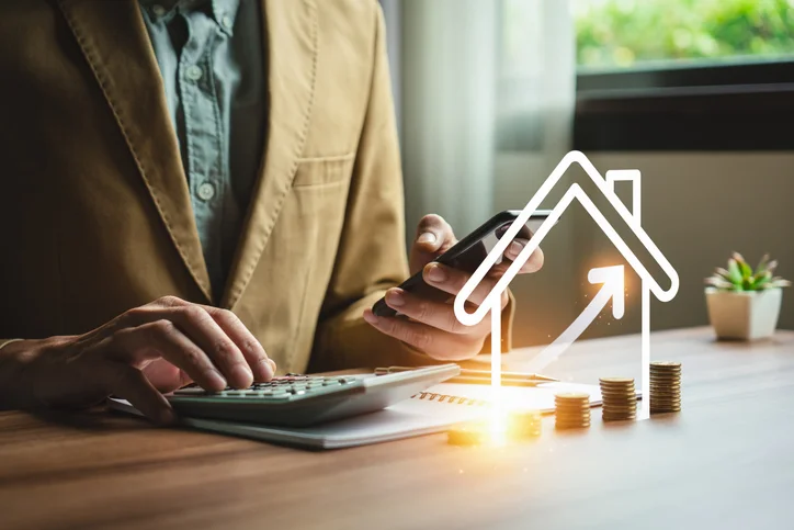 Homme tenant un smartphone dans une main et une calculatrice dans l'autre devant des piles de pièces de monnaie et un hologramme de maison