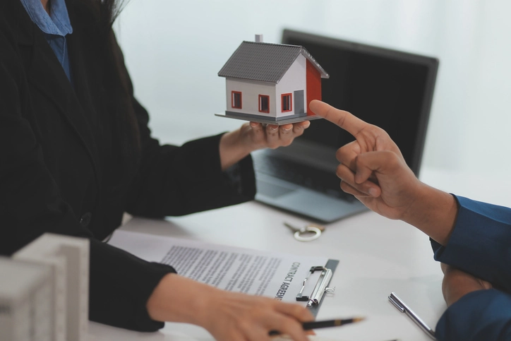 Main qui pointe une petite maison tenue par une autre personne, des documents, clé et ordinateur portable en arrière-plan