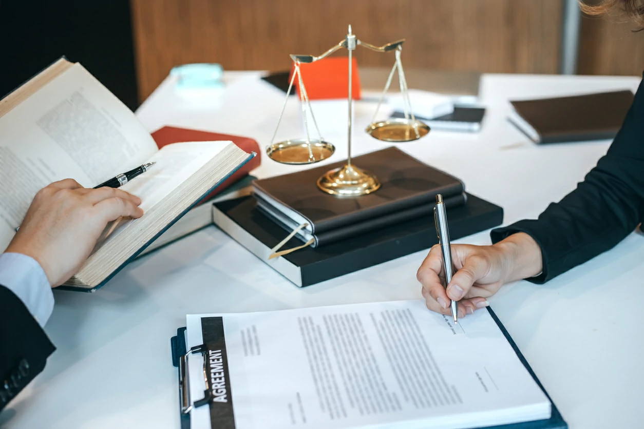 Homme tenant un livre et un stylo devant un professionnel juridique qui signe un document.