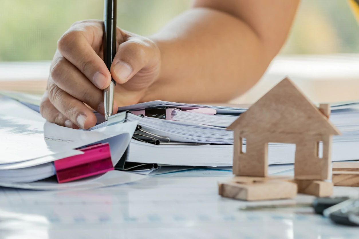 Main d'une femme en train de signer un document à coté d'une petite maison en bois.