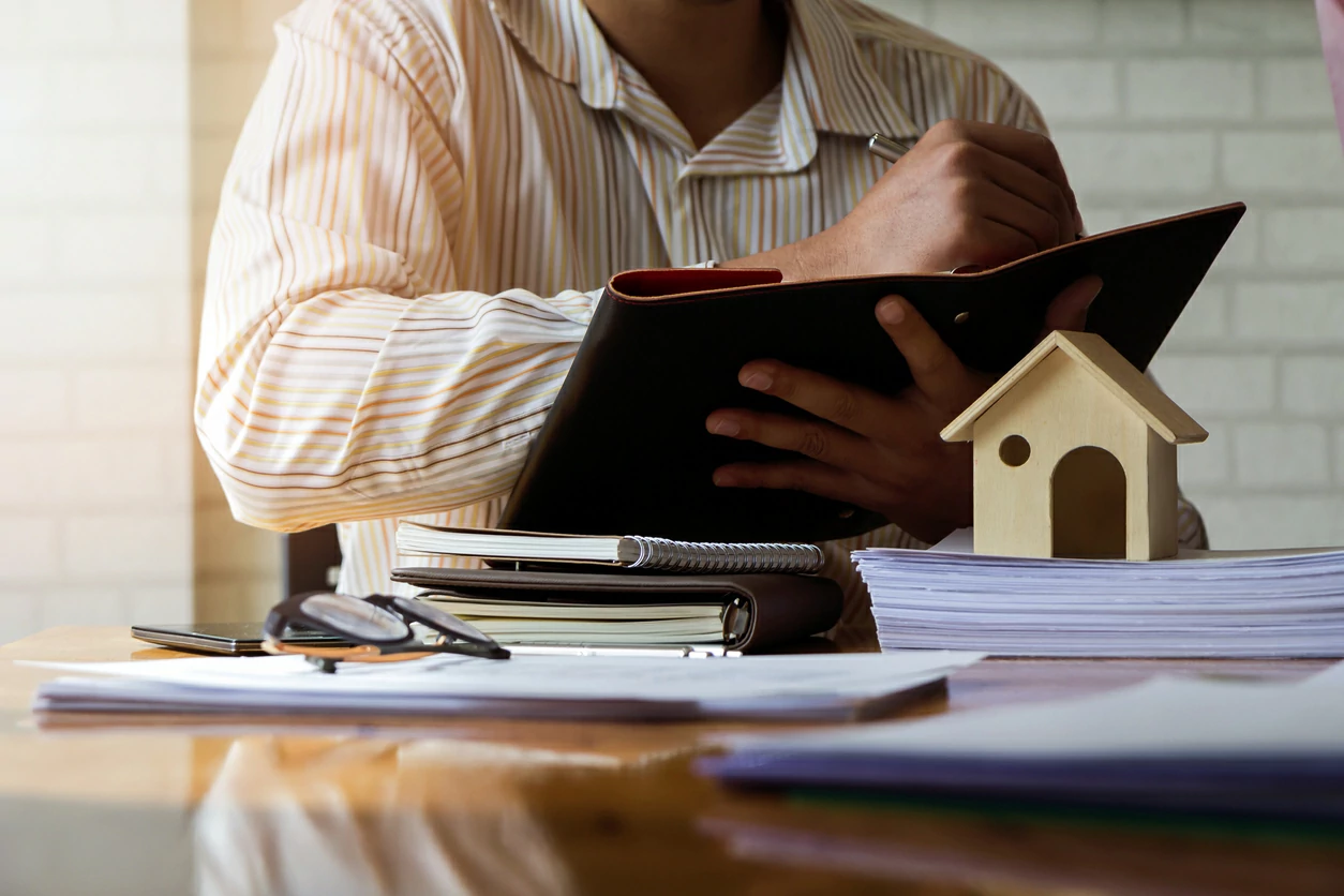 Homme en train de signer un document au dessus d'une petite maison en bois et d'autres documents éparpillés.