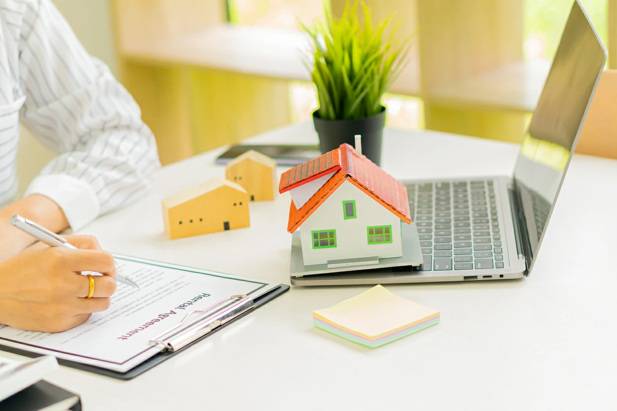 Personne signant un document coté d'un ordinateur portable et une petite maison.