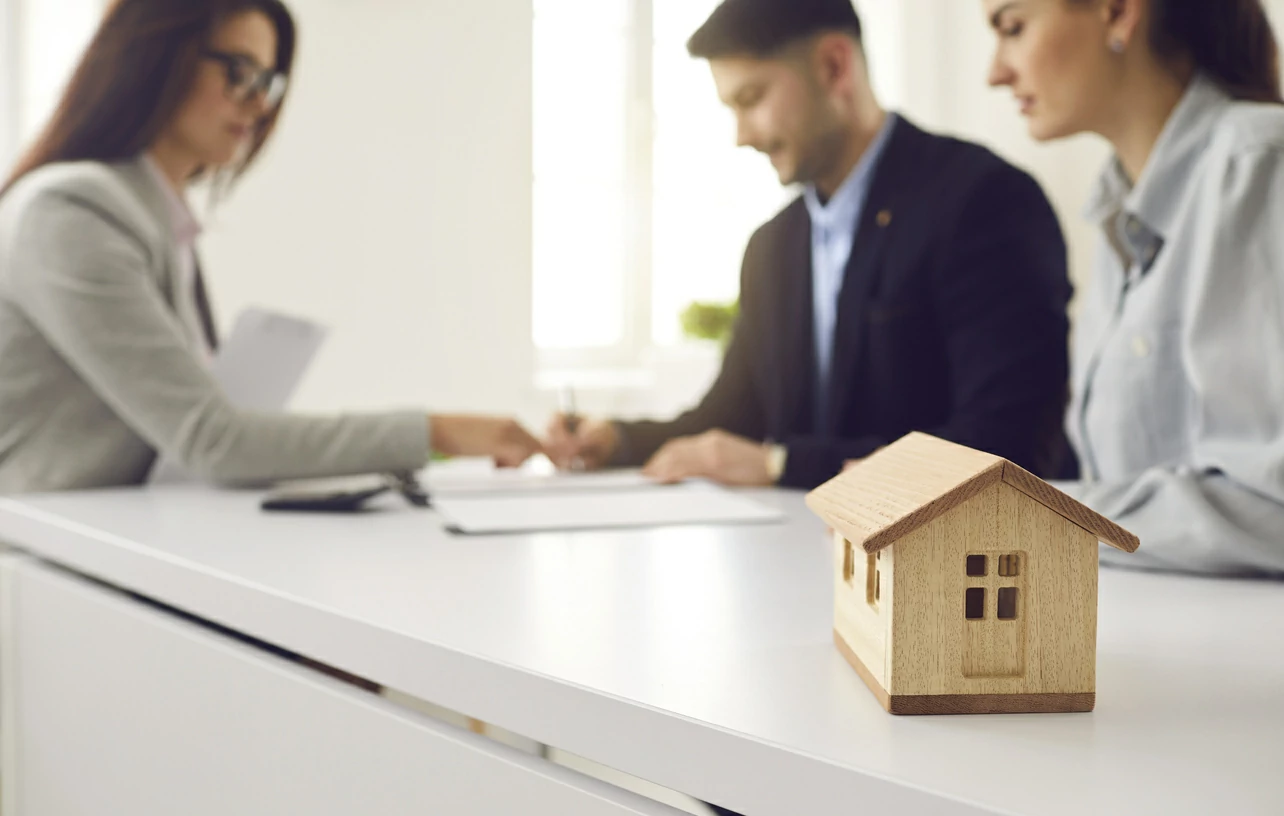 Petite maison en bois et trois personnes dont un homme qui signe un document en arrière plan.