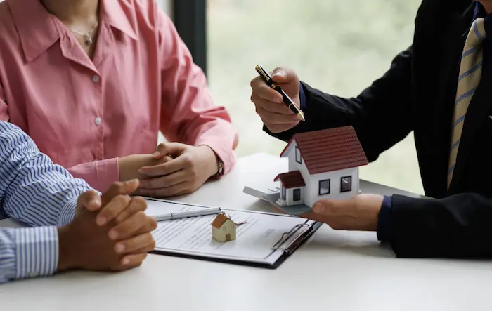 Homme pointant une petite maison dans sa main avec un stylo devant un couple et divers documents.