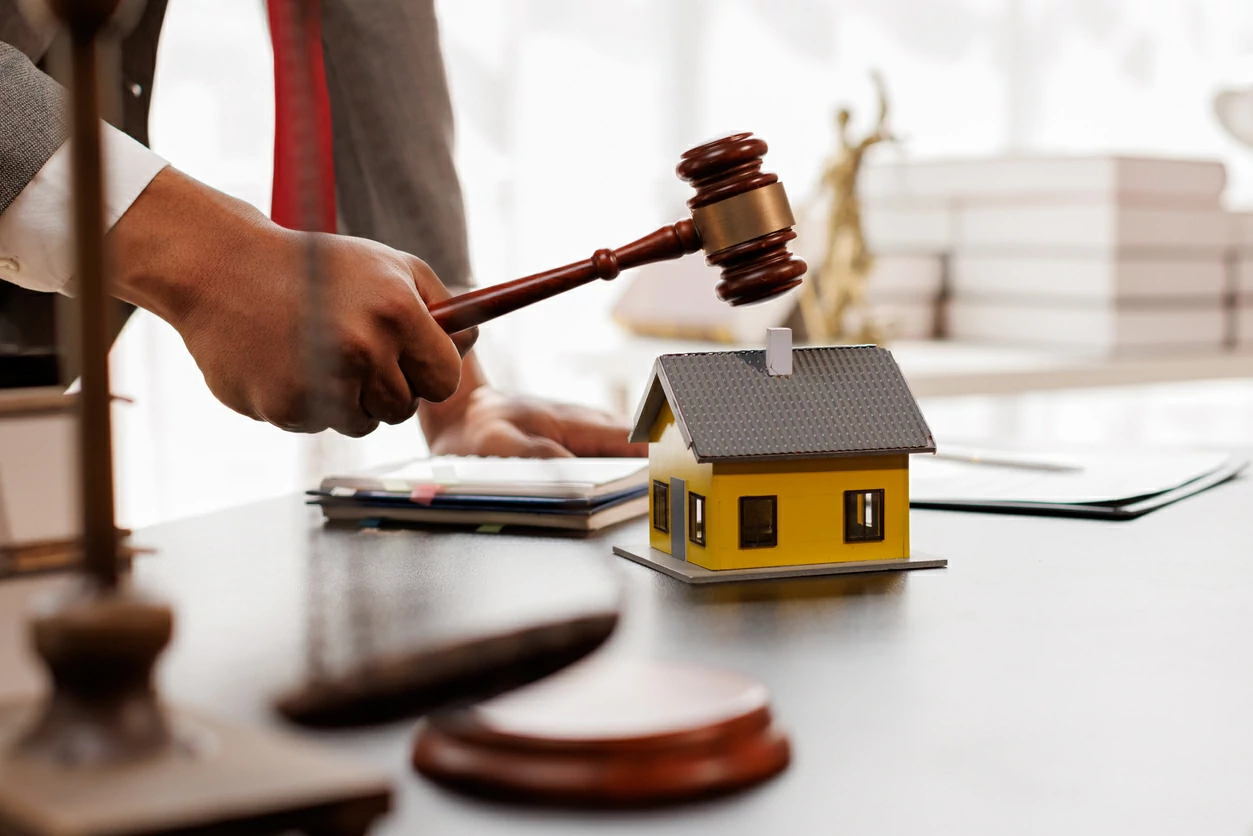 Main tenant un marteau en bois à coté d'une petite maison, divers documents et une balance de justice.c