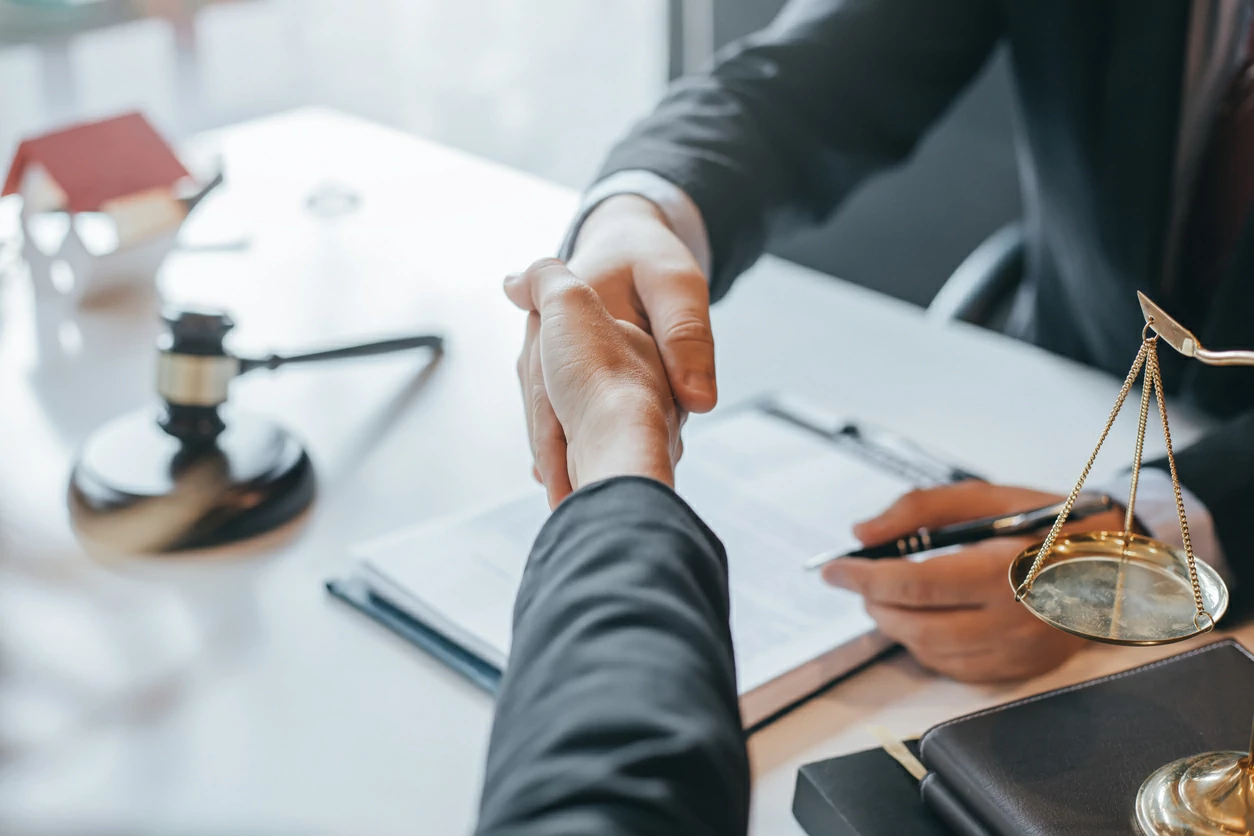 Poignée de main au-dessus d'un document, un marteau en bois, une petite balance de justice et une petite maison en arrière-plan.