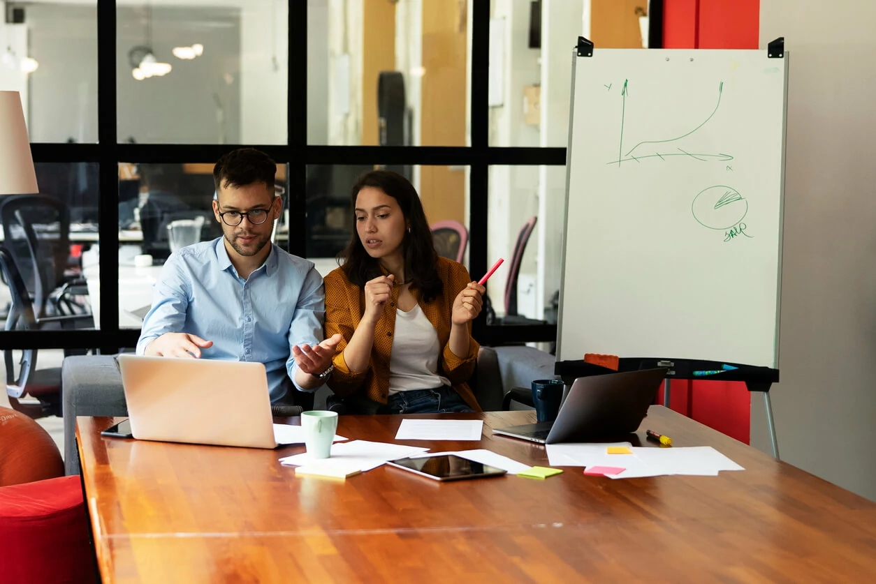 Un homme et une femme devant des documents et des ordinateurs et un tableau blanc en arrière.