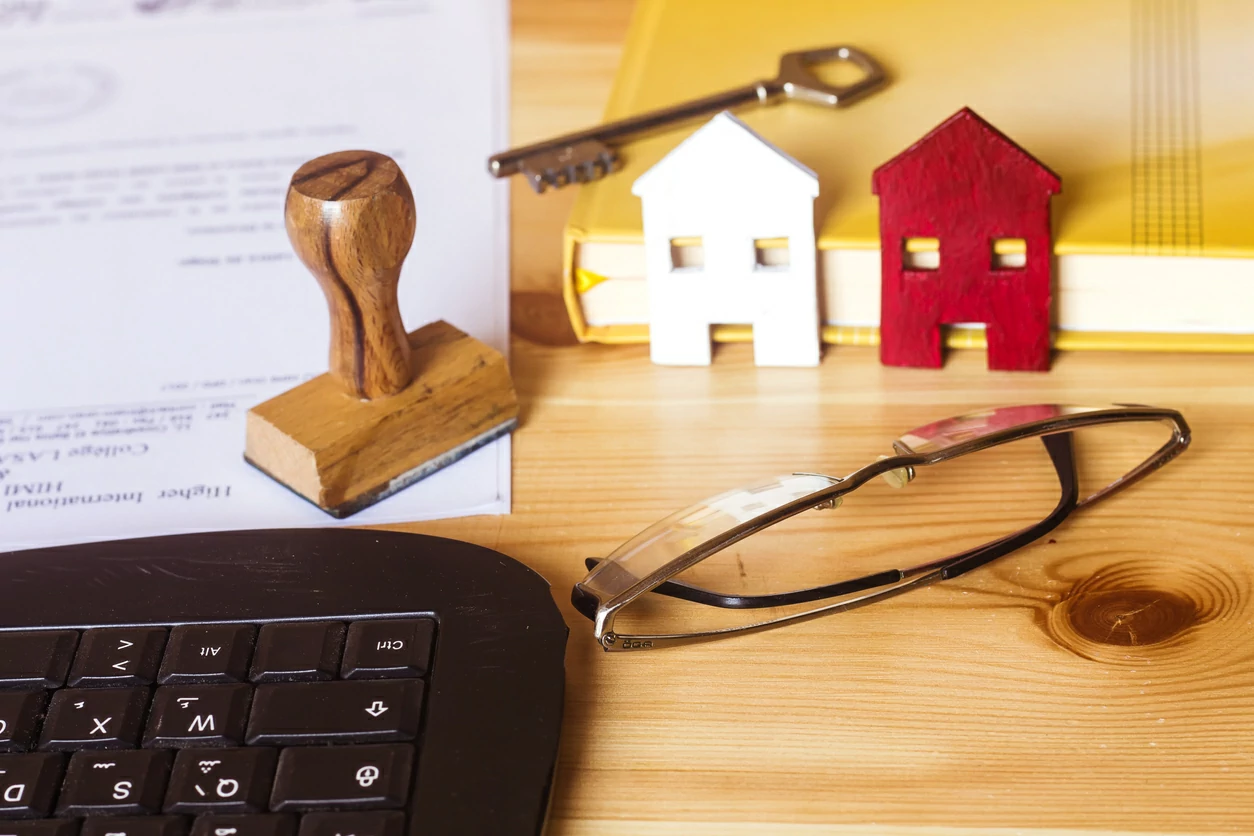 Clavier d'ordinateur, document avec tampon posé dessus, des lunettes, deux petites maison en papier et une clé.