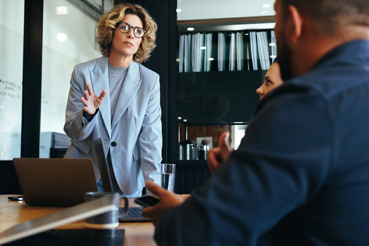 Femme debout devant un homme et une femme autour d'une table remplie de documents.