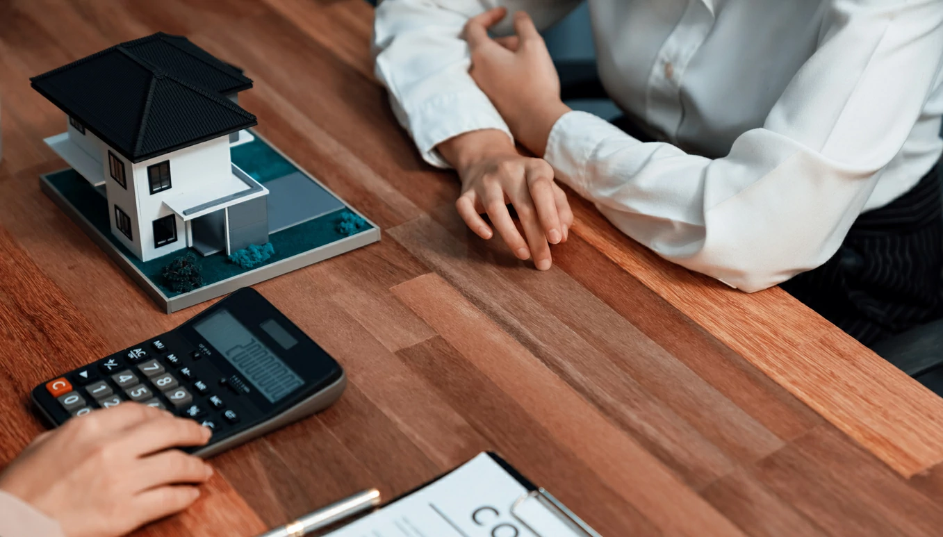 Main utilisant une calculatrice devant des documents, une maquette de maison et une femme.