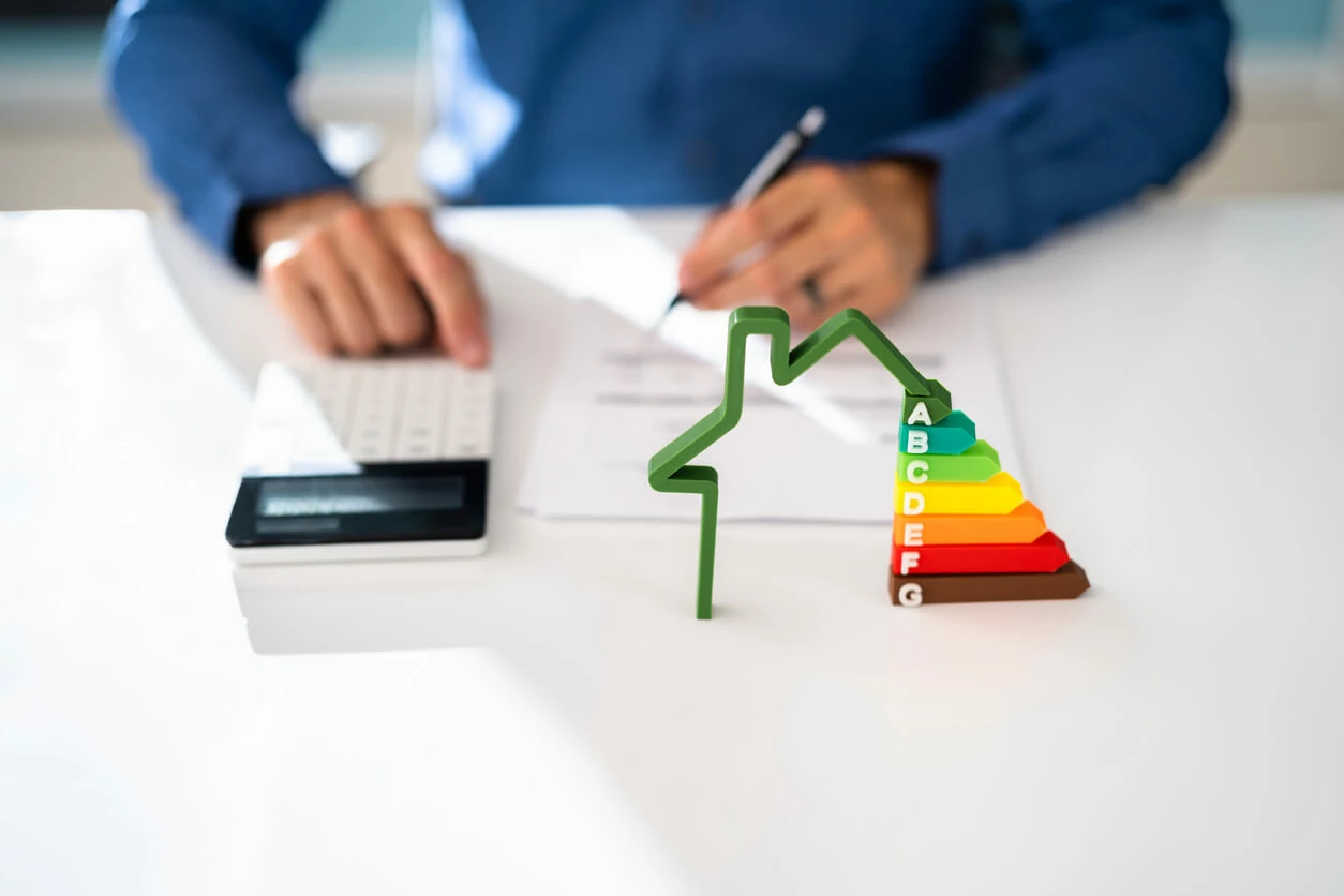 Main écrivant sur un document et un utilisant une calculatrice à coté d'une maison avec des classements énergétiques.