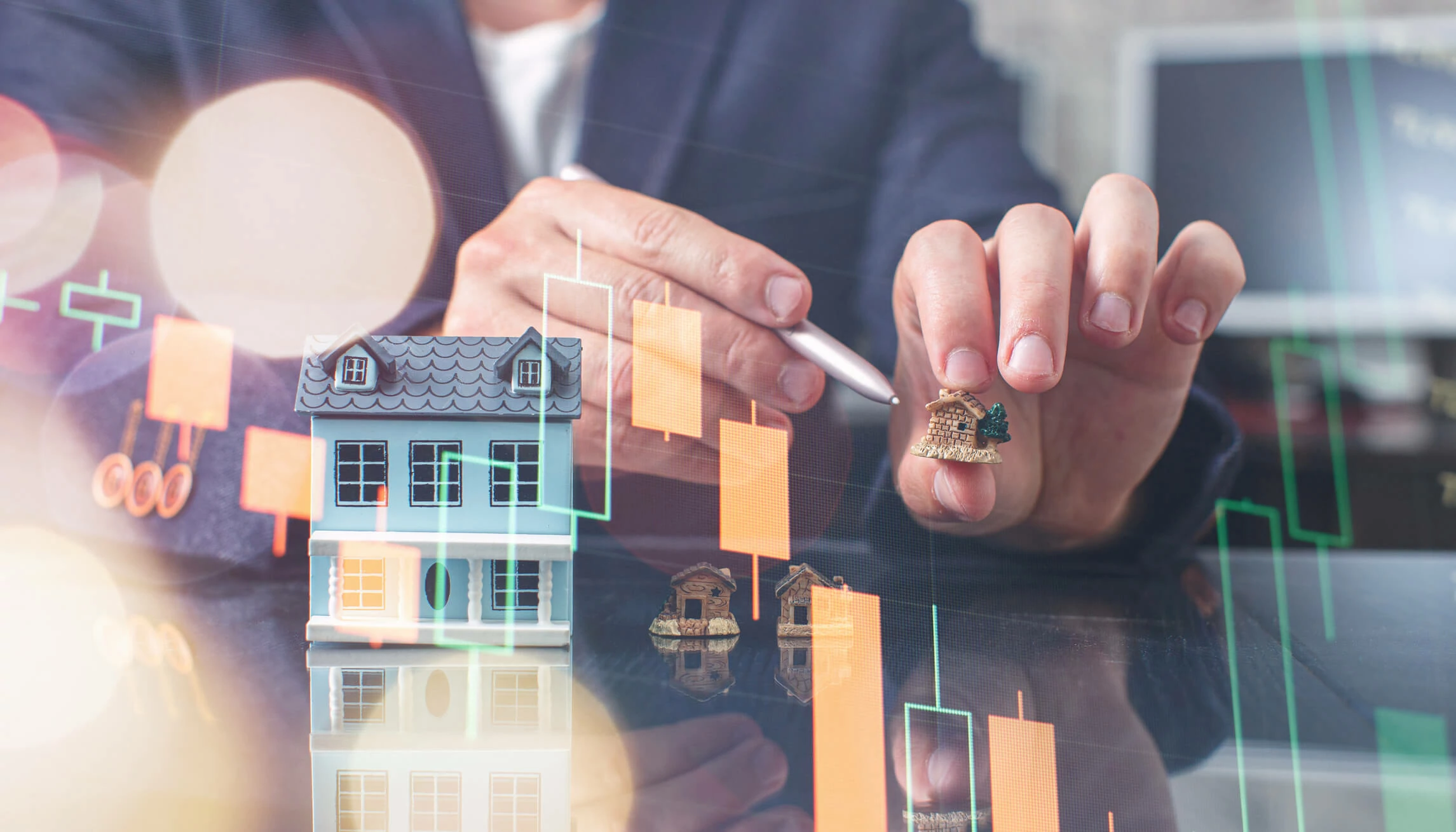 Main tenant une maison miniature et un stylo à coté d'une maquette de maison et d'une graphique de bougie japonaise baissière.